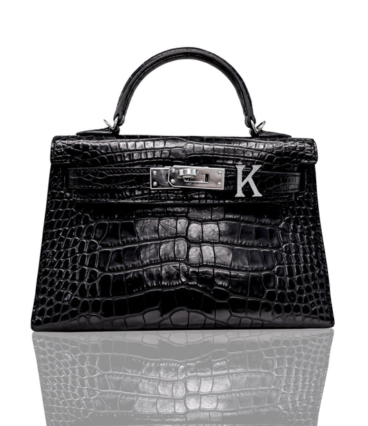 A black handbag with a crocodile pattern and a metallic letter 'K' emblem attached to the top, collection-preview.