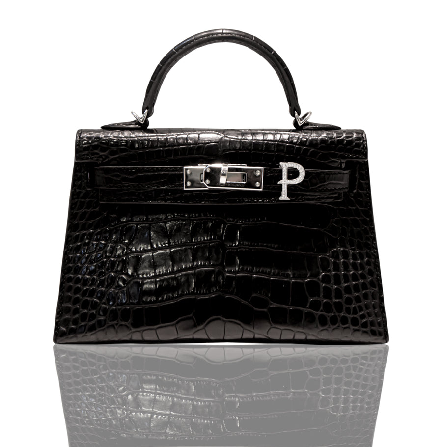A black handbag with a crocodile pattern and a silver initial 'P' brooch attached to it, collection-preview.