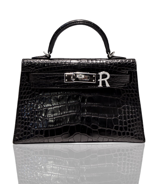 A black handbag with a crocodile pattern and a silver brooch with the letter 'R' attached to it, collection-preview.